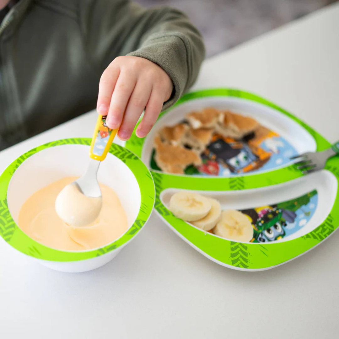 Child using a spoon with a bowl and plate featuring cartoon characters on a white surface.